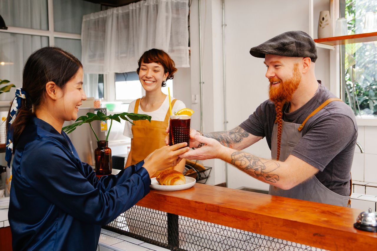 hero-img Glad hipster male employee passing glass of Black Velvet cocktail to smiling ethnic colleague at work